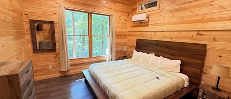 Habitació d'una cabana de fusta amb un llit, una petita arca, una finestra amb cortines i un aparell d'aire condicionat.