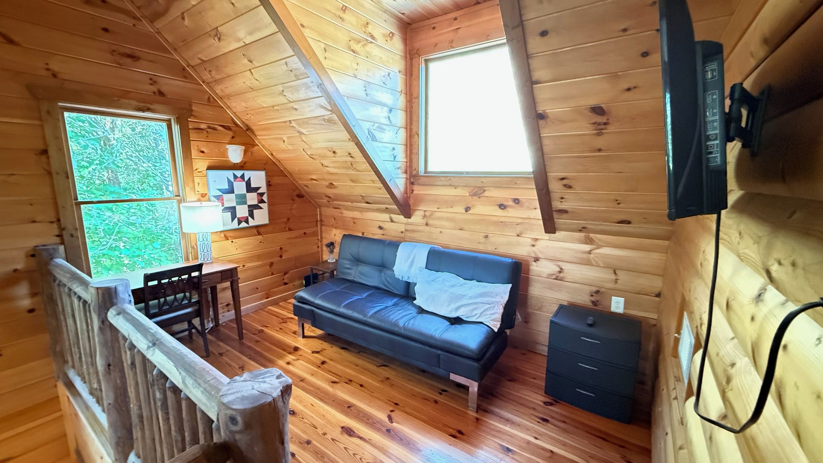 A cozy cabin with a loft bedroom, a wooden floor, a sofa, a desk, and a window with a view of trees.