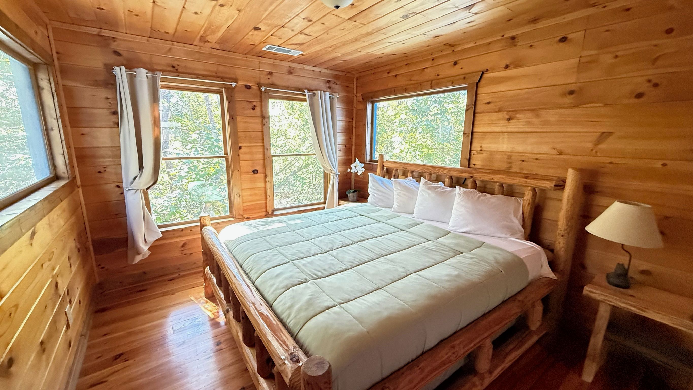 A wooden cabin bedroom with a bed, curtains, a nightstand, and a lamp.