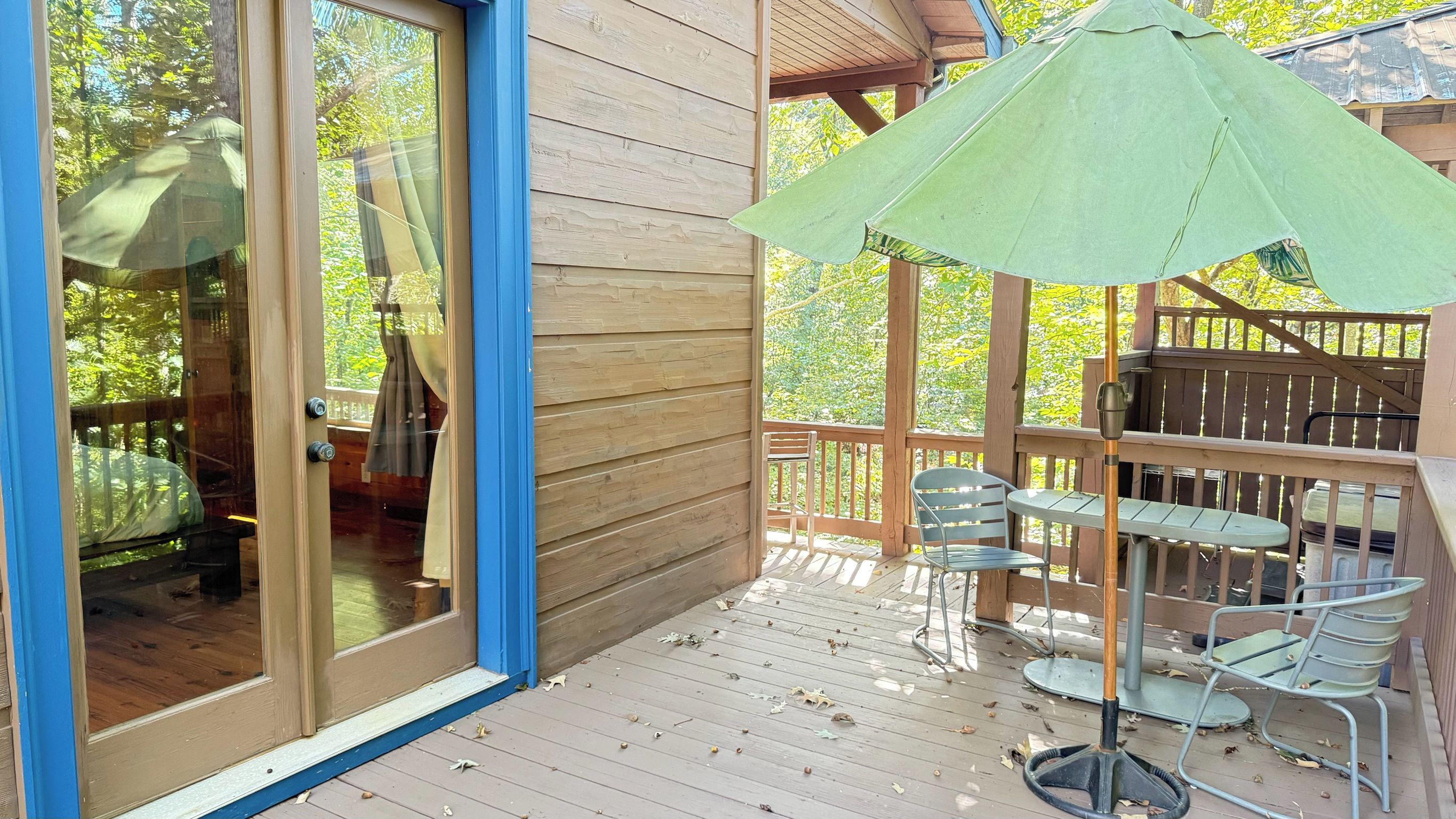 Een houten terras met een tafel en stoelen, een groene parasol en uitzicht op de omliggende bomen.