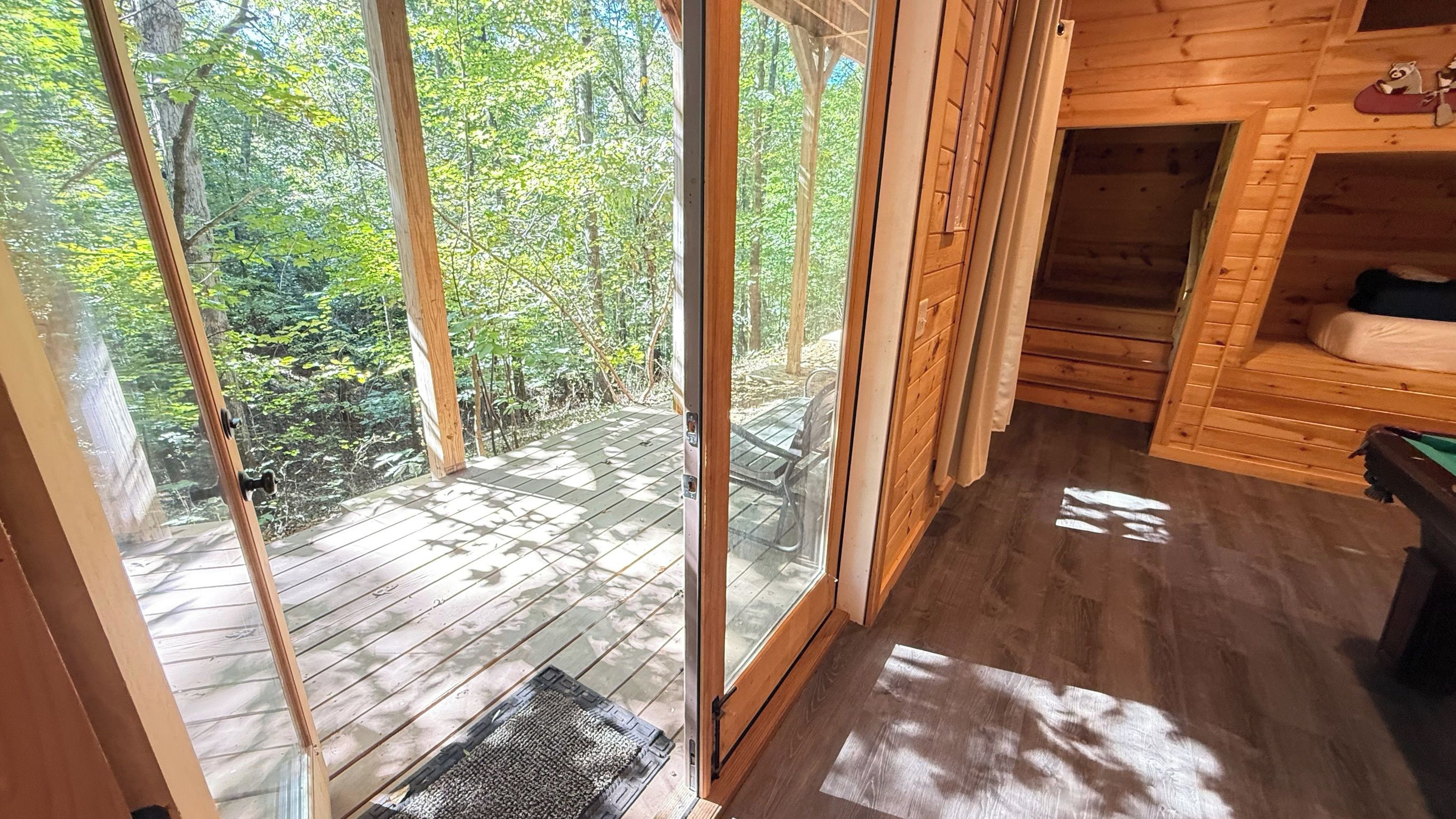 A wooden cabin with a glass door leading to a deck, a cozy interior with a bed and a small table, and a forested backdrop.