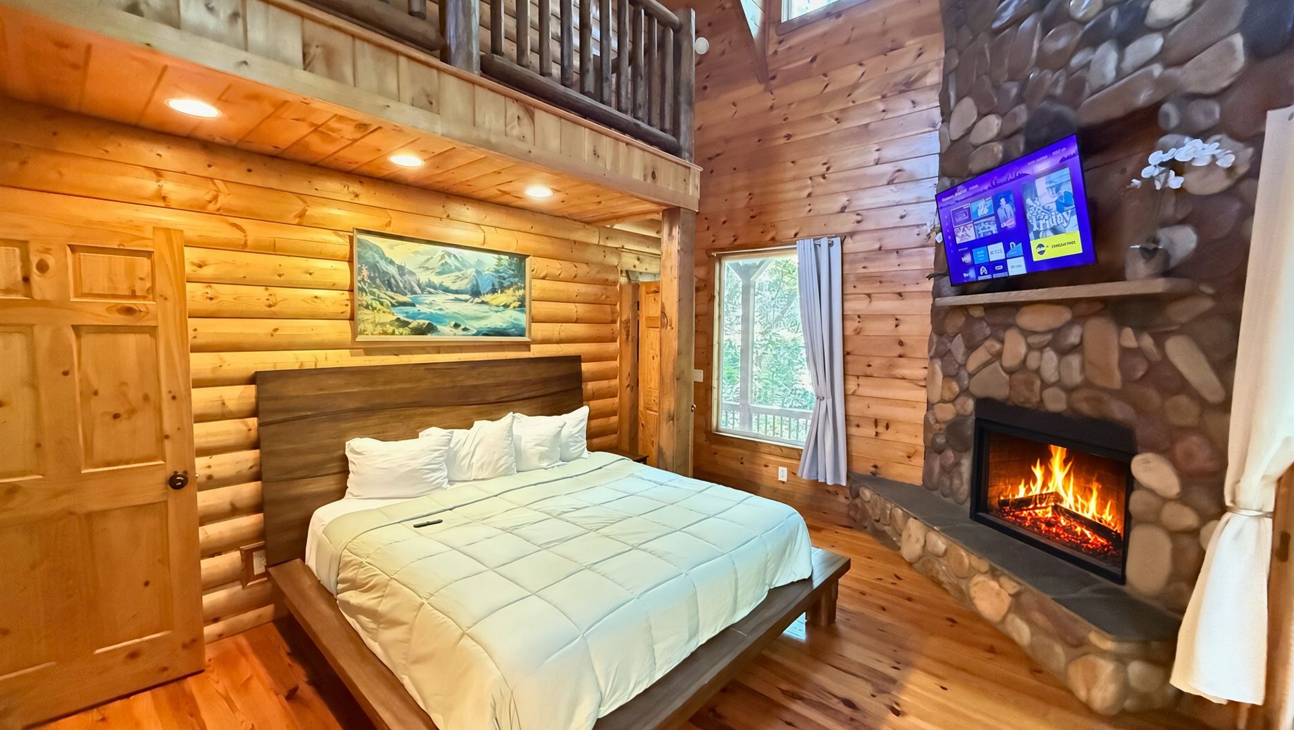 A log cabin bedroom with a bed, a stone fireplace, a TV on a shelf, and a painting on the wall.