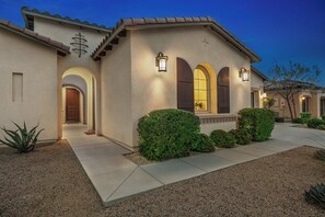 A stucco building with a tiled roof, a front entrance with a door, and a small garden area with shrubs and a cactus. - Resort-Style Escape with Pool, Spa, BBQ & Fire Pit (La Quinta)