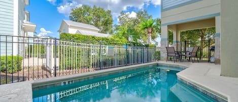 A rectangular swimming pool with a metal fence, surrounded by a patio area with chairs and a grill.