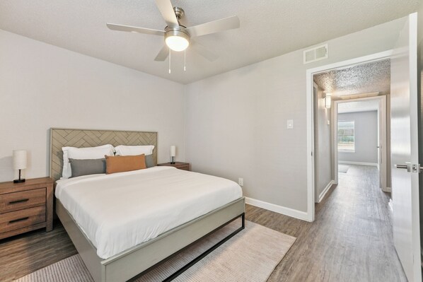 A bedroom with a bed, nightstands, a ceiling fan, and a door leading to another room.