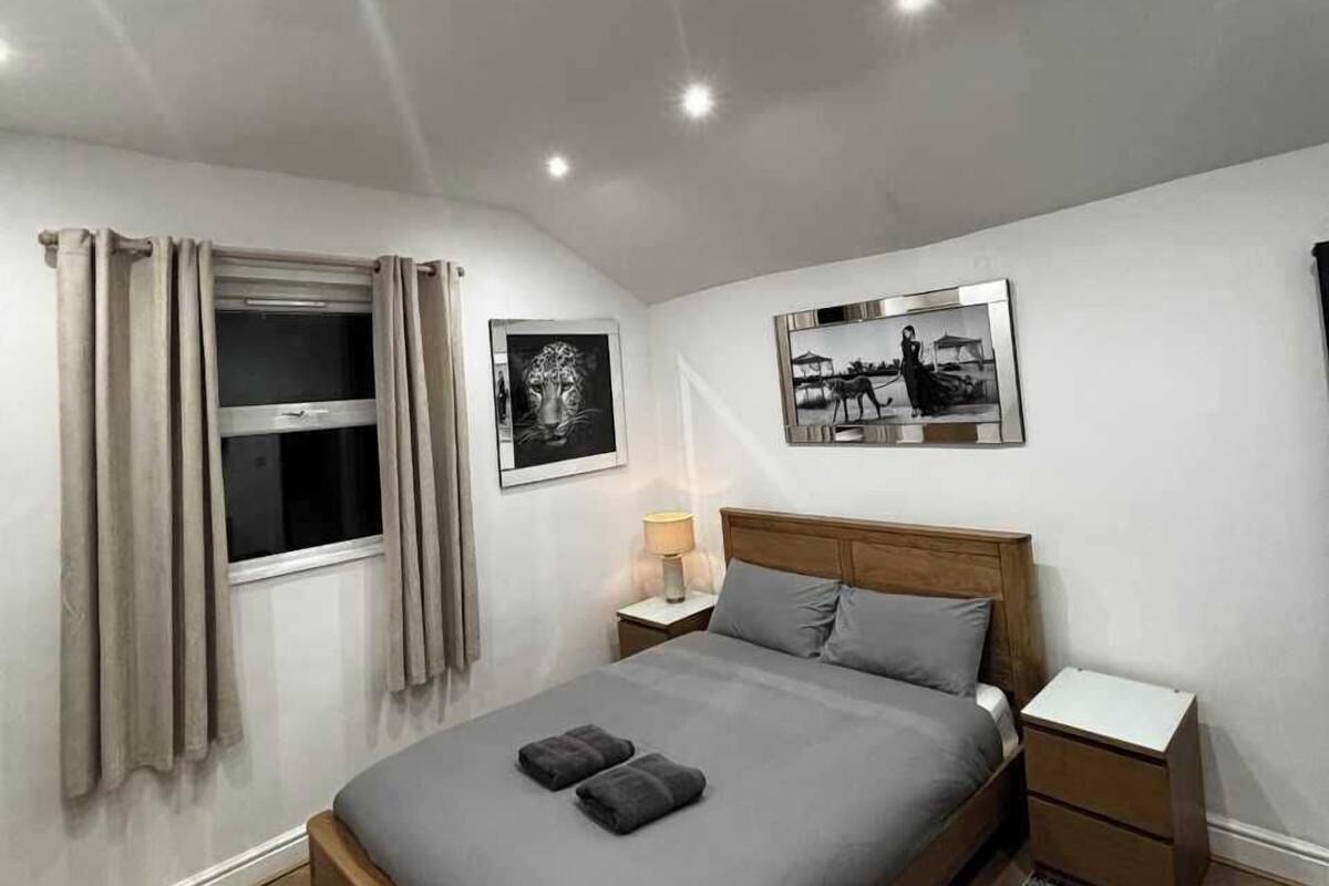 Un dormitorio moderno con una cama grande, una mesita de noche, dos cuadros enmarcados en la pared y una ventana con cortinas.