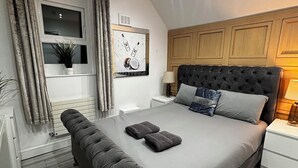 Un dormitorio moderno con una cama grande, cabecera, mesita de noche y cortinas en la ventana. Incluye una obra de arte colgada en la pared.