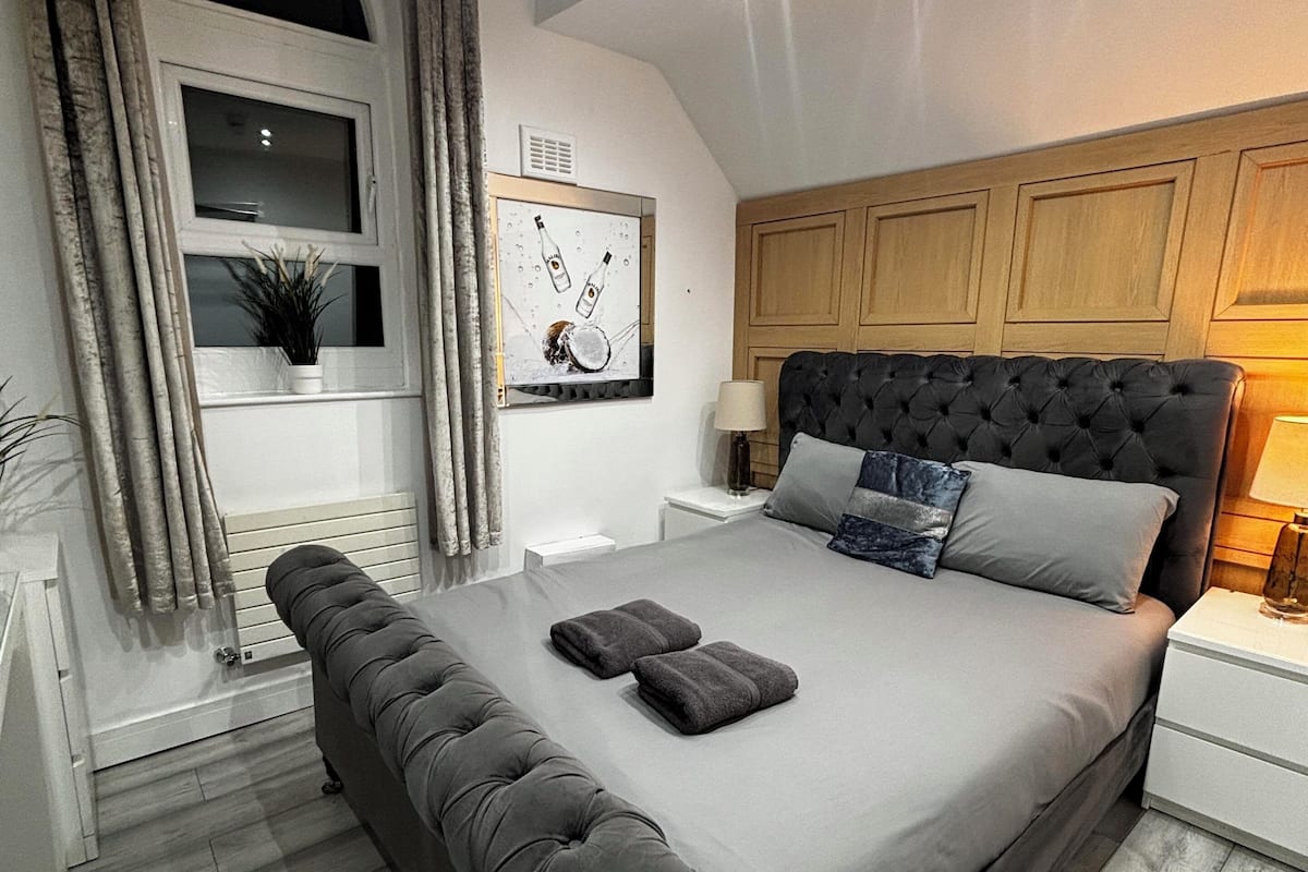 Un dormitorio moderno con una cama grande, cabecera, mesita de noche y cortinas en la ventana. Incluye una obra de arte colgada en la pared.