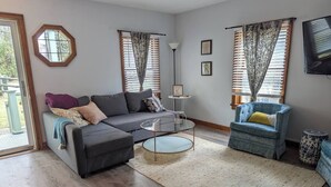 Ein Wohnzimmer mit einem grauen Ecksofa, einem runden Couchtisch, einem blauen Sessel und einem großen Fenster mit gemusterten Vorhängen.