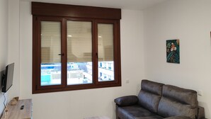 Een woonkamer met een bruine leren bank, een houten salontafel, een televisie op een dressoir, en een raam met zonwering.