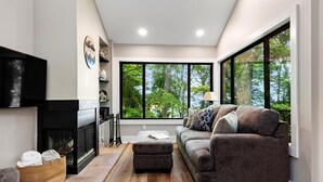 A modern living room with a fireplace, a sofa, a coffee table, and large windows offering a view of trees. - Dobie 24 with Wintergreen Advantage (Wintergreen)