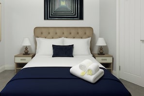A neatly made bed with a dark blue bedspread, two white pillows, and a dark blue accent pillow. There are two bedside tables with lamps, and a wall art piece above the bed.