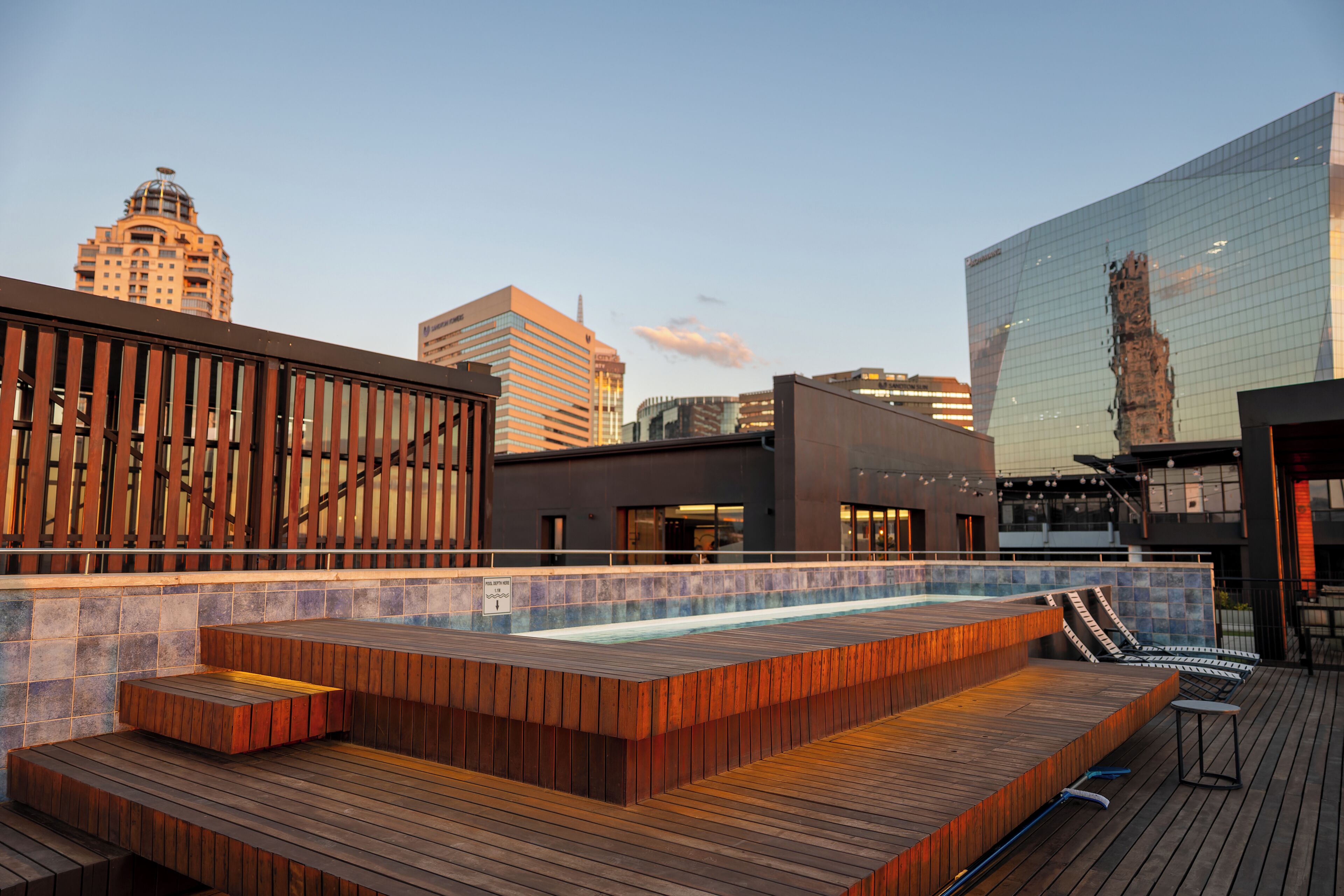 Zona de piscina en la azotea con escalones de madera y sillones de descanso, rodeada de edificios modernos y un cielo despejado.