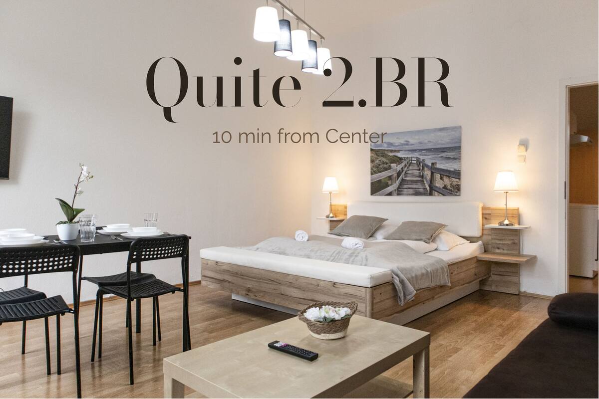 A modern bedroom with a wooden bed, a dining table with chairs, a small table with a remote control, and a wall-mounted artwork.