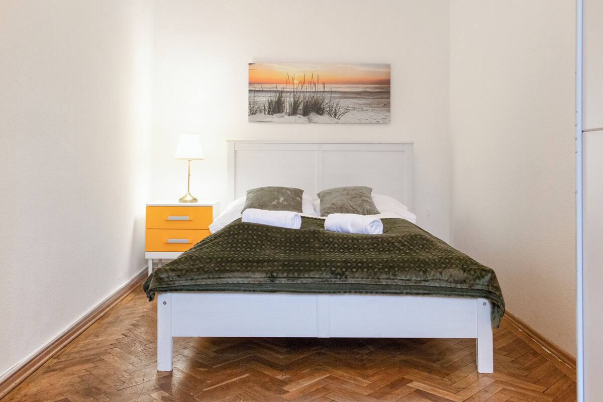 A neatly made bed with a patterned blanket, a bedside table with a lamp, and a wall-mounted painting of a sunset over a beach.