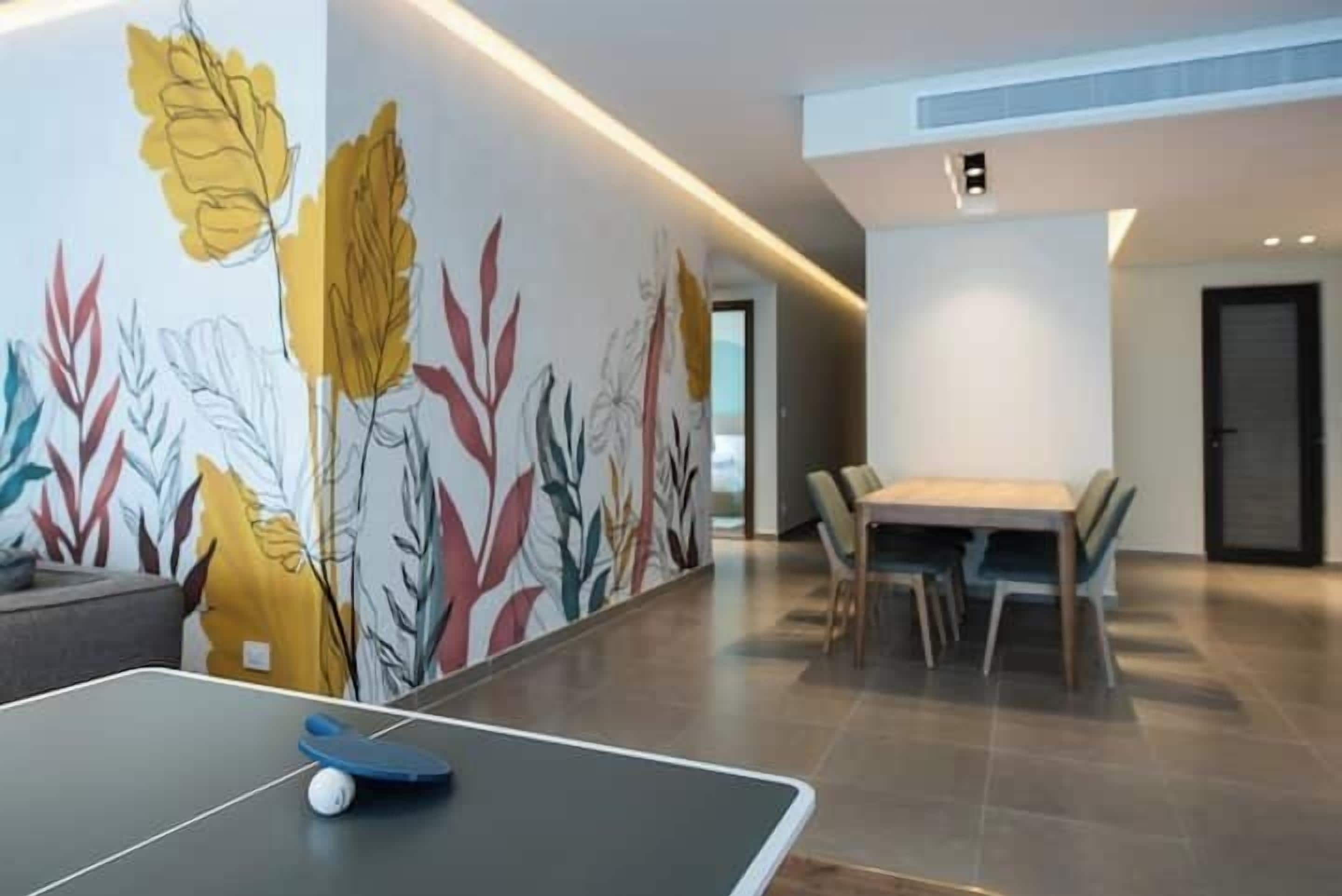 A modern living room with a ping pong table, a dining area with a wooden table and chairs, and a colorful wall mural.