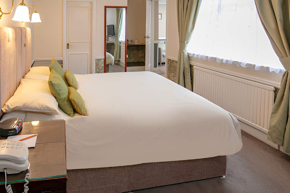 A hotel room with a large bed, bedside table with a telephone and a remote control, a mirror, and a window with curtains.