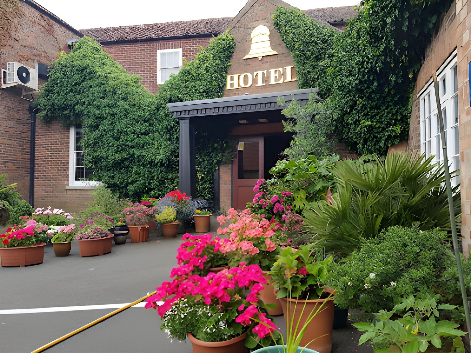 A hotel with a traditional brick facade, a bell-shaped sign, and a variety of potted plants and flowers in the entrance area.