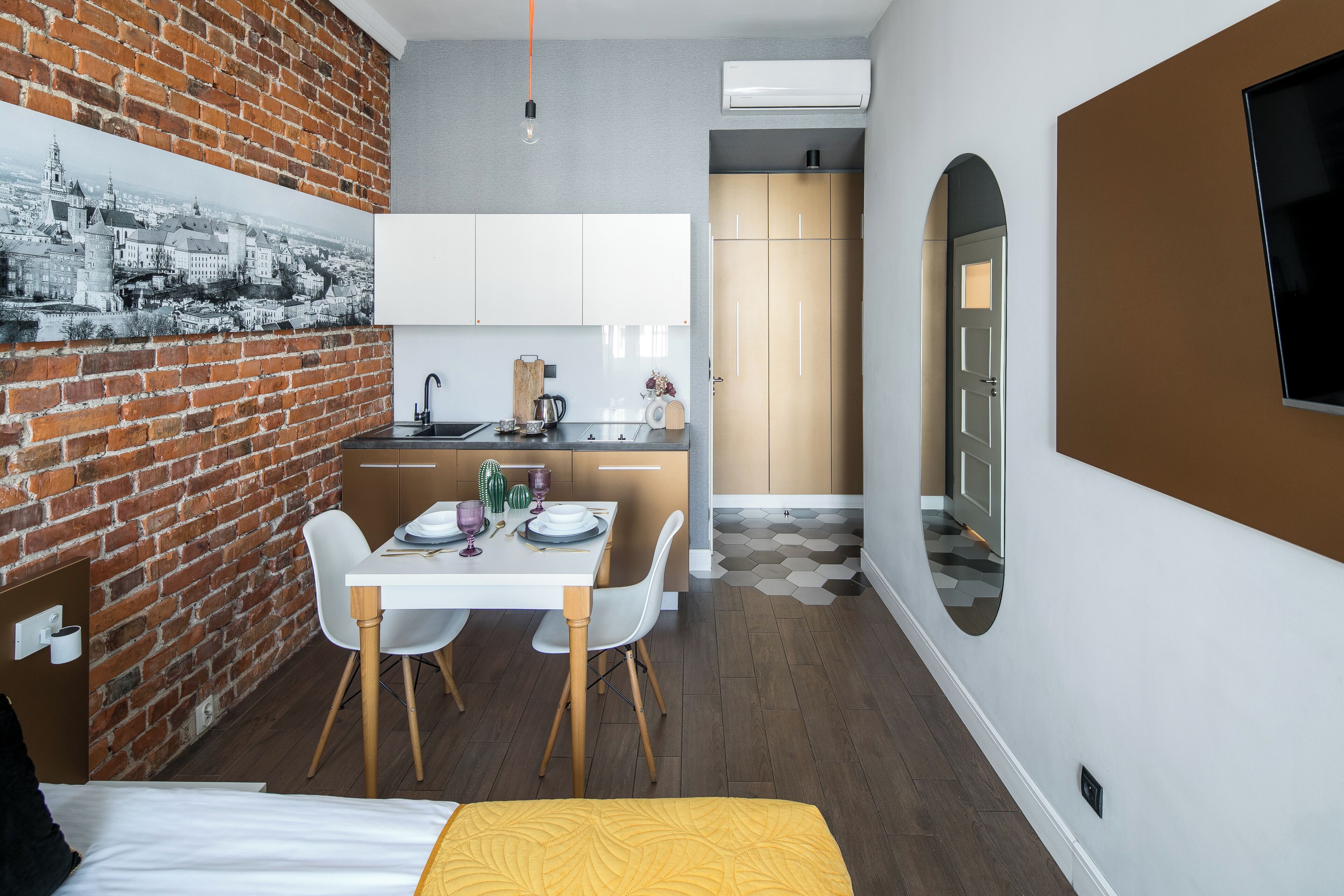 A compact hotel room with a bed, a dining table with chairs, a kitchen area with cabinets and a sink, and a large window.