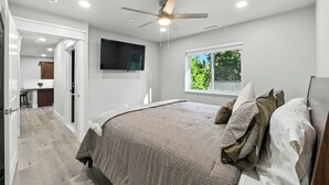 Une chambre à coucher avec un lit, un ventilateur de plafond, un téléviseur fixé au mur et une fenêtre donnant sur un paysage verdoyant.