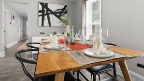 A dining area with a wooden table set for four, surrounded by black chairs. There is a large abstract painting on the wall, a window with blinds, and a plant on the windowsill.