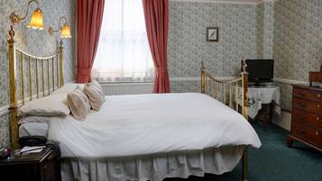 A neatly made bed with a white duvet, a wooden nightstand with a telephone, a desk with a television, and a dresser.