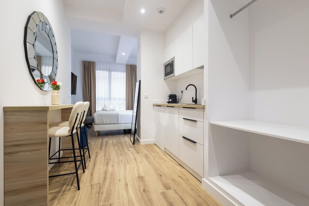 Une chambre d’hôtel compacte comprenant un coin cuisine, un lit, un espace salon avec un fauteuil et un grand miroir.