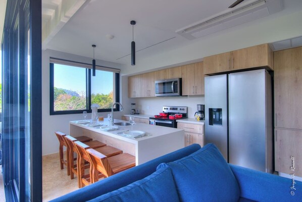 Cozinha moderna com ilha central, armários de madeira, geladeira de aço inoxidável e área de jantar com sofá azul.