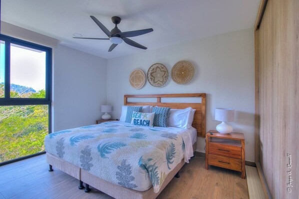A bedroom with a bed, a ceiling fan, bedside tables, and a view of greenery outside. - Great views apartment with King bed Torres del Mar (Jacó, Torres del Mar)