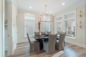 A dining room with a dark wooden table and six chairs, a chandelier, and large windows with blinds.