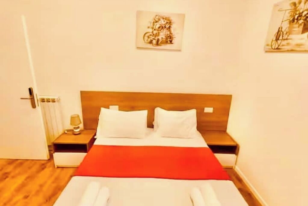 Habitació d'hotel amb capçal de llit de fusta, estribo vermell i almoina blanca. Hi ha una nitòria amb una làmpada i una taula petita.
