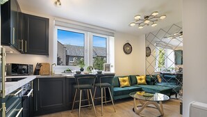 A modern kitchen with dark cabinetry, a central island, bar stools, and a green sofa in a bright living area with a view of houses. - Jackson Terrace HL6 - Grampian Lettings (Aberdeen)