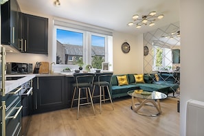 A modern kitchen with dark cabinetry, a central island, bar stools, and a green sofa in a living area with a view of houses.