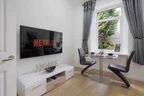 A modern living room with a flat-screen TV displaying 'NETFLIX,' a glass-top dining table with chairs, a white entertainment unit with 'HOME' sign, and a window with curtains.
