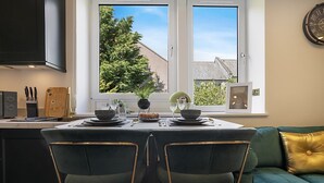 Uma cozinha moderna com uma mesa de jantar posta para dois, uma janela com vista para árvores e edifícios, e um sofá verde com almofada dourada.