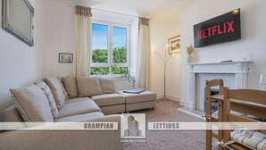A living room with a sofa, a fireplace, a television, and a window with curtains. - Abbey Place Apartment | Grampian Lettings (Aberdeen)