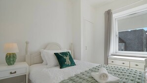 A neatly arranged bedroom with a bed, pillows, a bedside table with a lamp, a window with curtains, and a view of buildings outside. - Abbey Place Apartment | Grampian Lettings (Aberdeen)