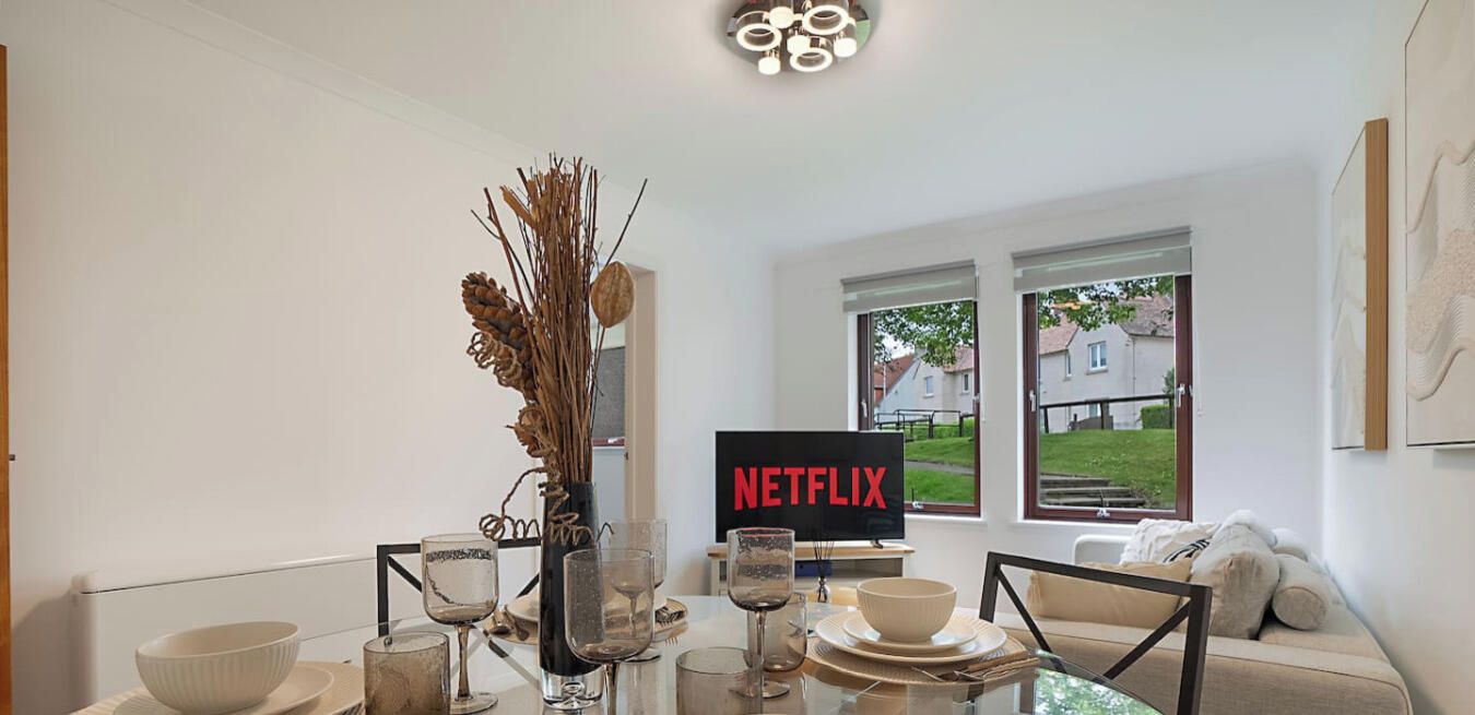 Una sala de estar moderna con una mesa de vidrio para cuatro personas, un sofá y un televisor que muestra el logotipo de Netflix.