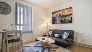 Una sala de estar con un sofá, una mesa redonda para café, una mesa de comedor con sillas, un reloj de pared y un cuadro de un paisaje urbano.