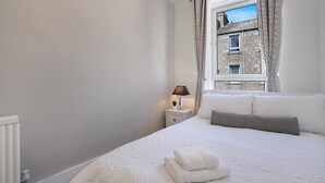 Un dormitorio con cama, una mesita de noche con lámpara, una ventana con cortinas y vistas a un edificio de piedra.