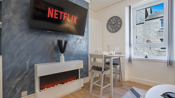 Una sala de estar moderna con chimenea, una mesa de comedor, un televisor montado en la pared y un reloj.