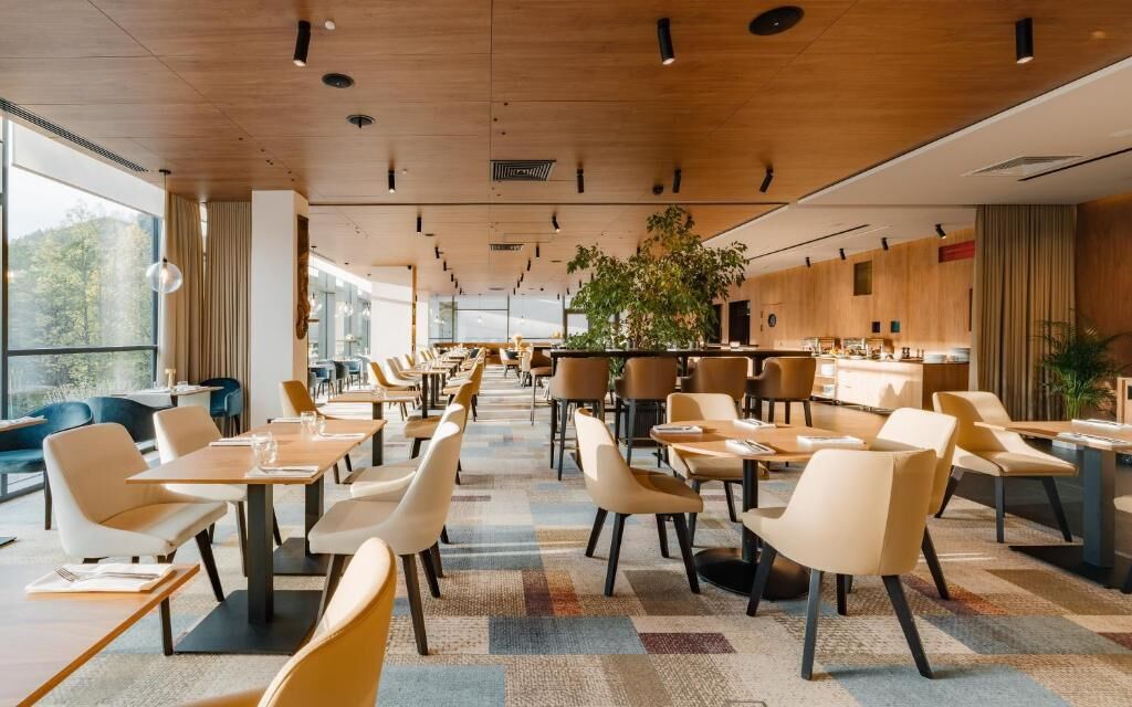 A modern restaurant with a long counter and multiple tables, large windows, and a wooden ceiling.