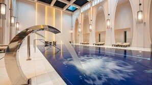 Una piscina interior moderna con un elemento acuático central, techo iluminado por el sol y asientos tipo lounge a lo largo de las paredes.
