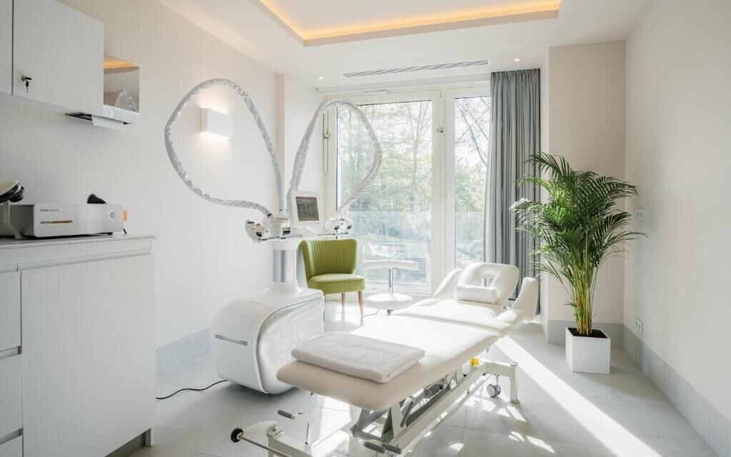 Een moderne behandelkamer met een bed, onderzoekstafel en een groene stoel.