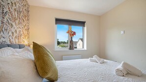 Ein ordentlich bezogenes Bett mit einem großen Kopfkissen, ein Fenster mit Blick auf Häuser und eine Heizung unter dem Fenster.
