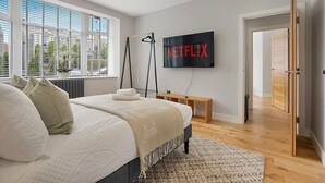 A modern bedroom with a large bed, a TV mounted on the wall, a window with blinds, and a view of greenery outside.