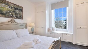 Un lit bien fait, avec une tête de lit, un coin fenêtre agrémenté de coussins et un tableau accroché au mur.
