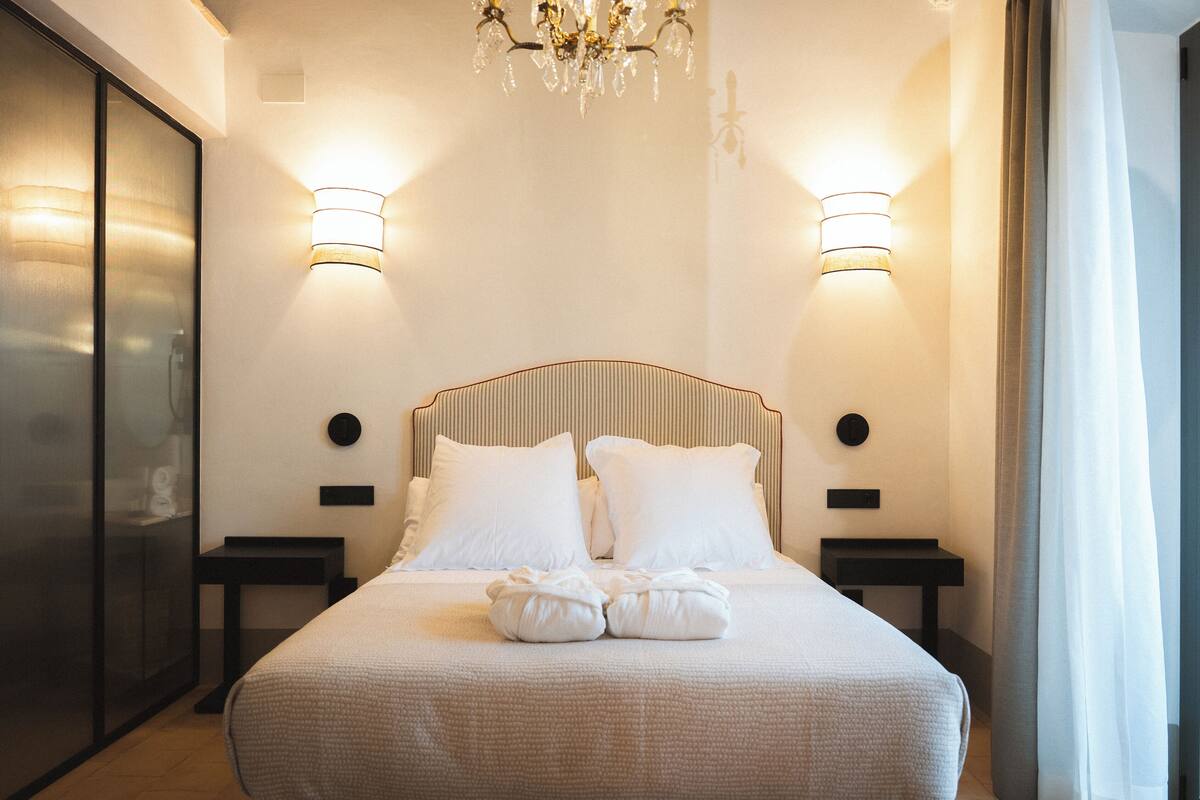 A neatly made bed with white linens, two bedside tables, a chandelier, and wall-mounted lighting.
