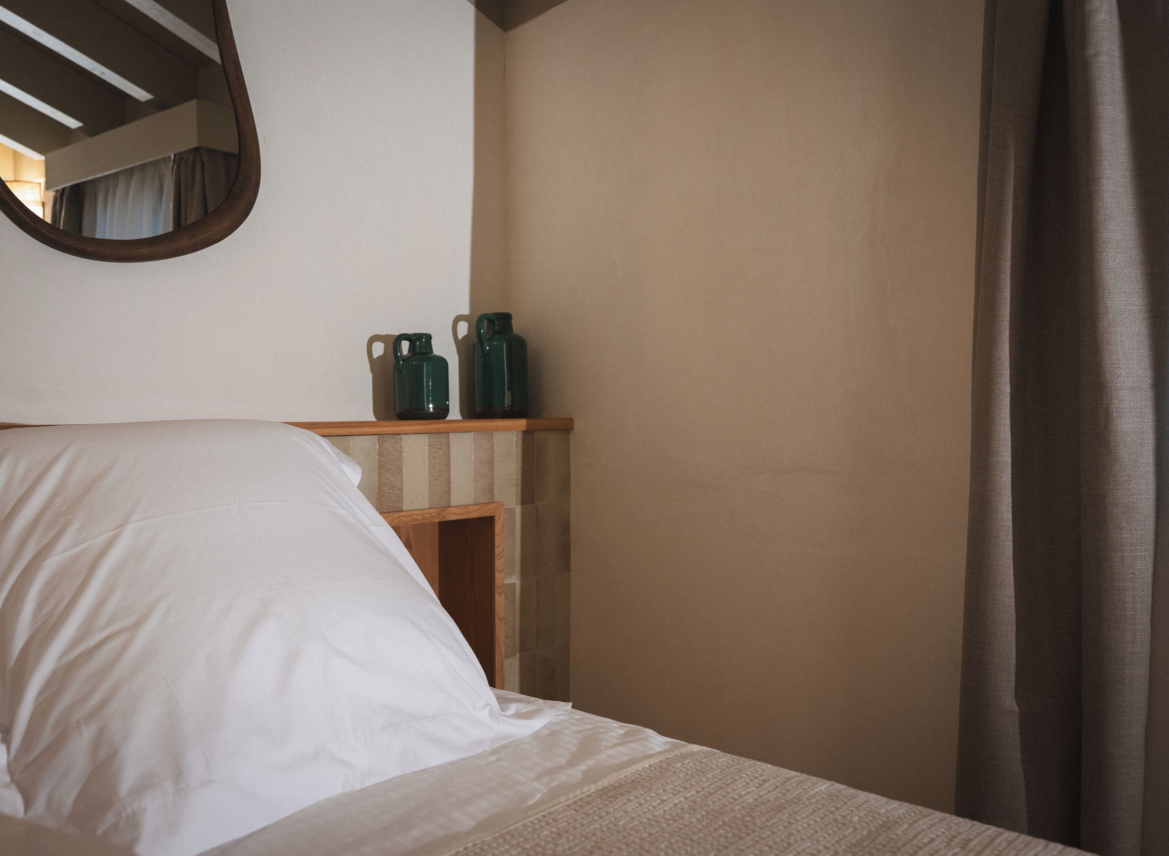 Un dormitorio con cama, una estantería de madera con dos jarras verdes y un espejo en la pared.