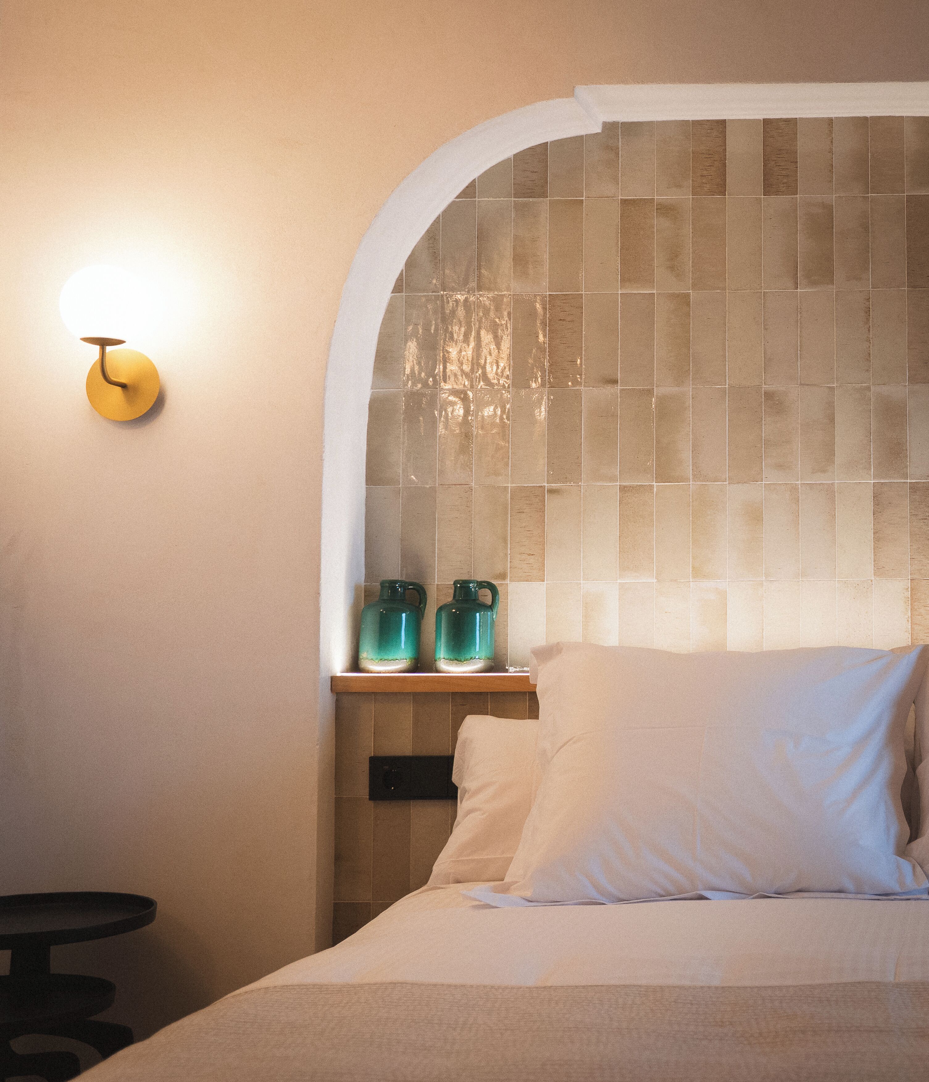 A bedroom with a bed, a nightstand, a wall-mounted light, and a shelf with two green bottles.